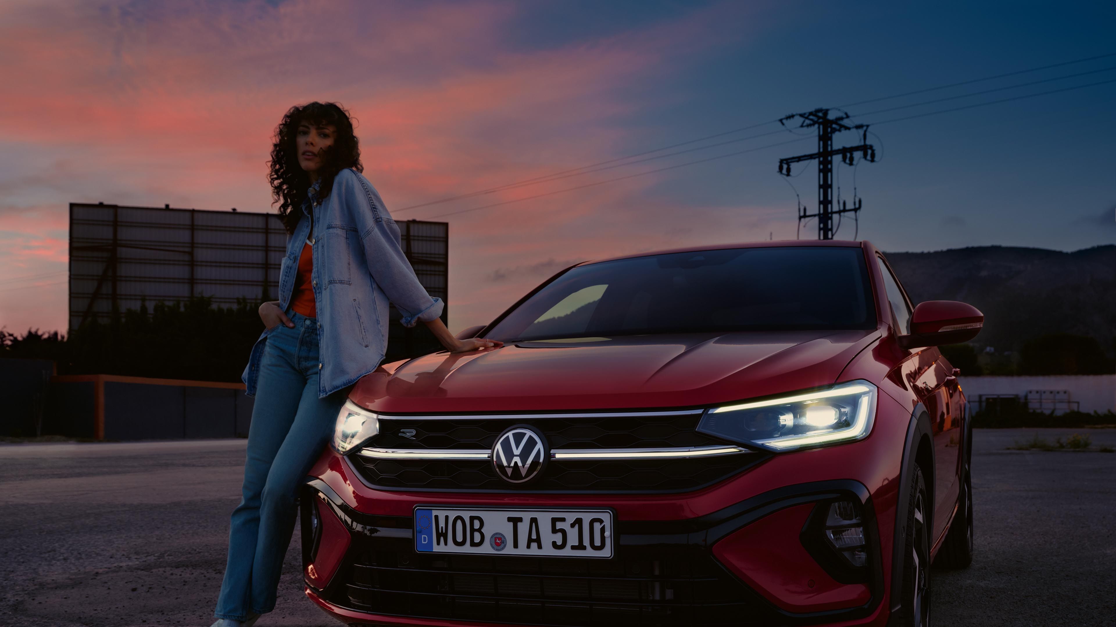 Rdeč VW Taigo R-Line na parkirišču. Luči so vključene. Ženska se naslanja na pokrov motornega prostora.