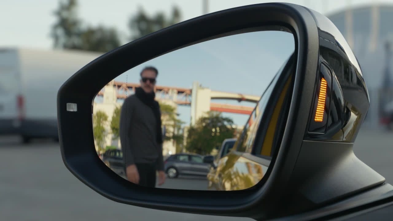 Strani vzvratni ogledalo avtomobila prikazuje osebo, ki stoji na parkirišču. Oseba je zamegljena, v ozadju pa je stavba in drevo. Vzvratni ogledalo prikazuje tudi rumeni signalni luč avtomobila.