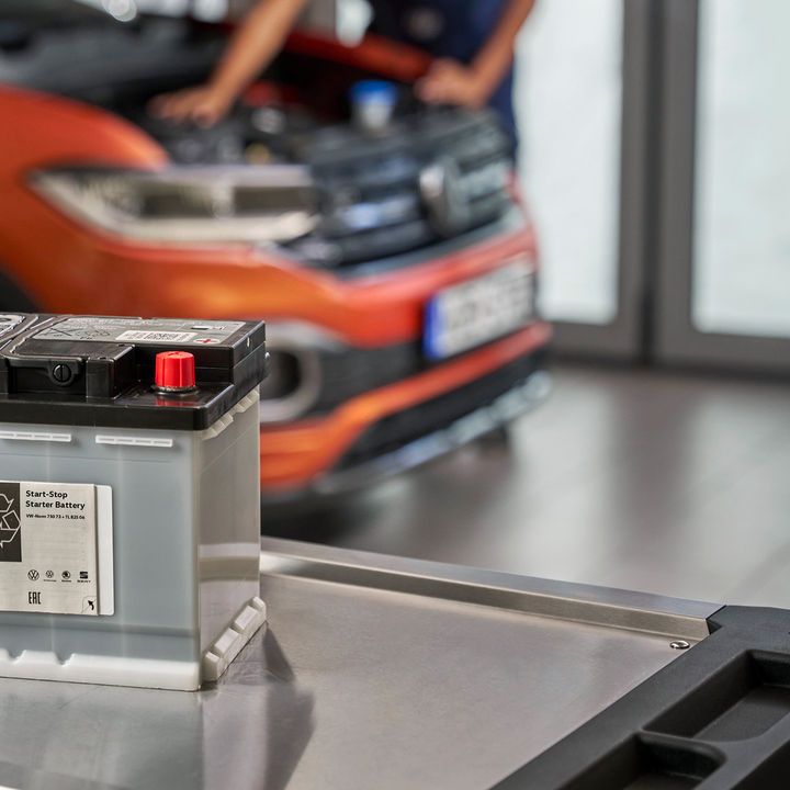 Akumulator za avto je postavljen na vozičku v mehanični delavnici. V ozadju je oranžen T-Cross z odprtim pokrovom motorja.