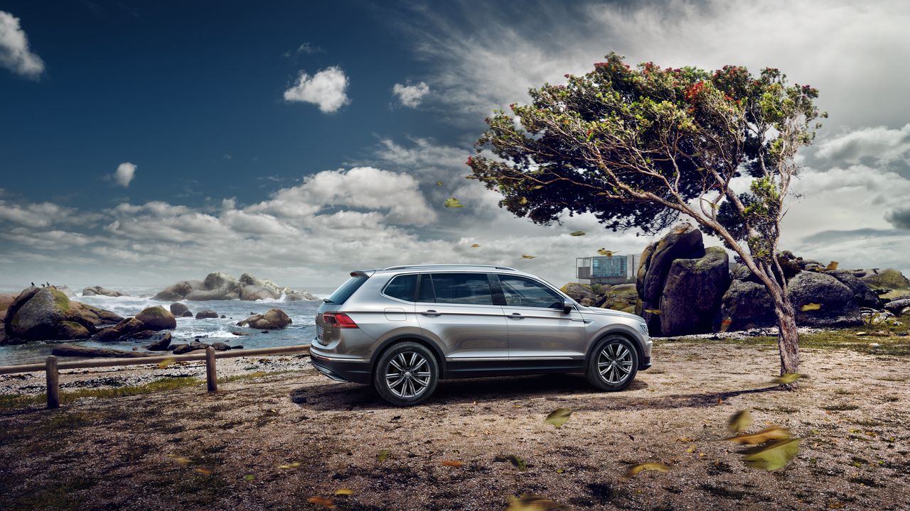 Srebrn VW Tiguan Allspace stoji ob obali, pod osamljenim drevesom. V ozadju so skalnati bregovi in morje pod oblačnim nebom. Listje na tleh nakazuje vetrovno vreme.