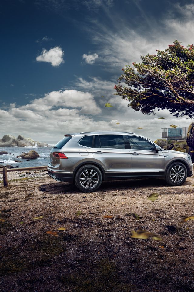 Srebrn VW Tiguan Allspace stoji ob obali, pod osamljenim drevesom. V ozadju so skalnati bregovi in morje pod oblačnim nebom. Listje na tleh nakazuje vetrovno vreme.