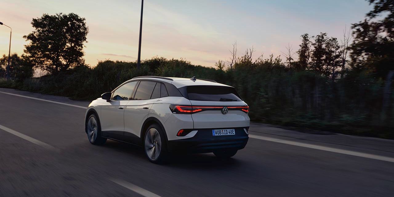 Od zadaj je prikazan bel VW ID.4, ki se ob sončnem zahodu pelje po regionalni cesti.