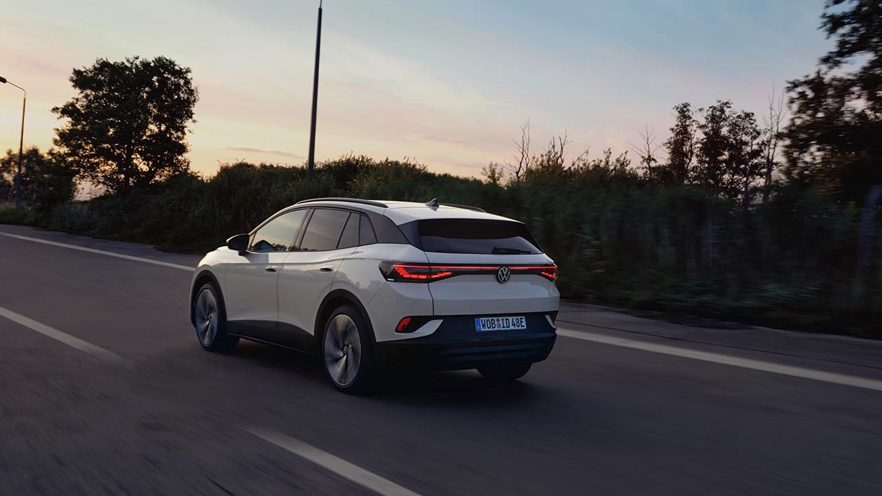Od zadaj je prikazan bel VW ID.4, ki se ob sončnem zahodu pelje po regionalni cesti.