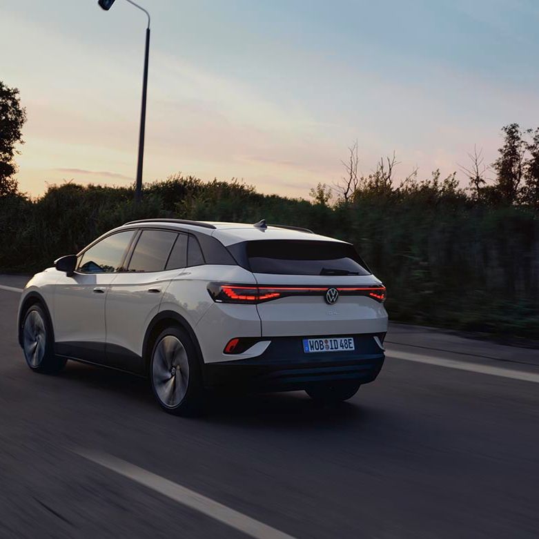 Od zadaj je prikazan bel VW ID.4, ki se ob sončnem zahodu pelje po regionalni cesti.