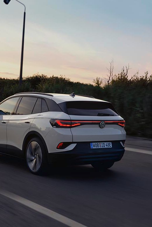 Od zadaj je prikazan bel VW ID.4, ki se ob sončnem zahodu pelje po regionalni cesti.