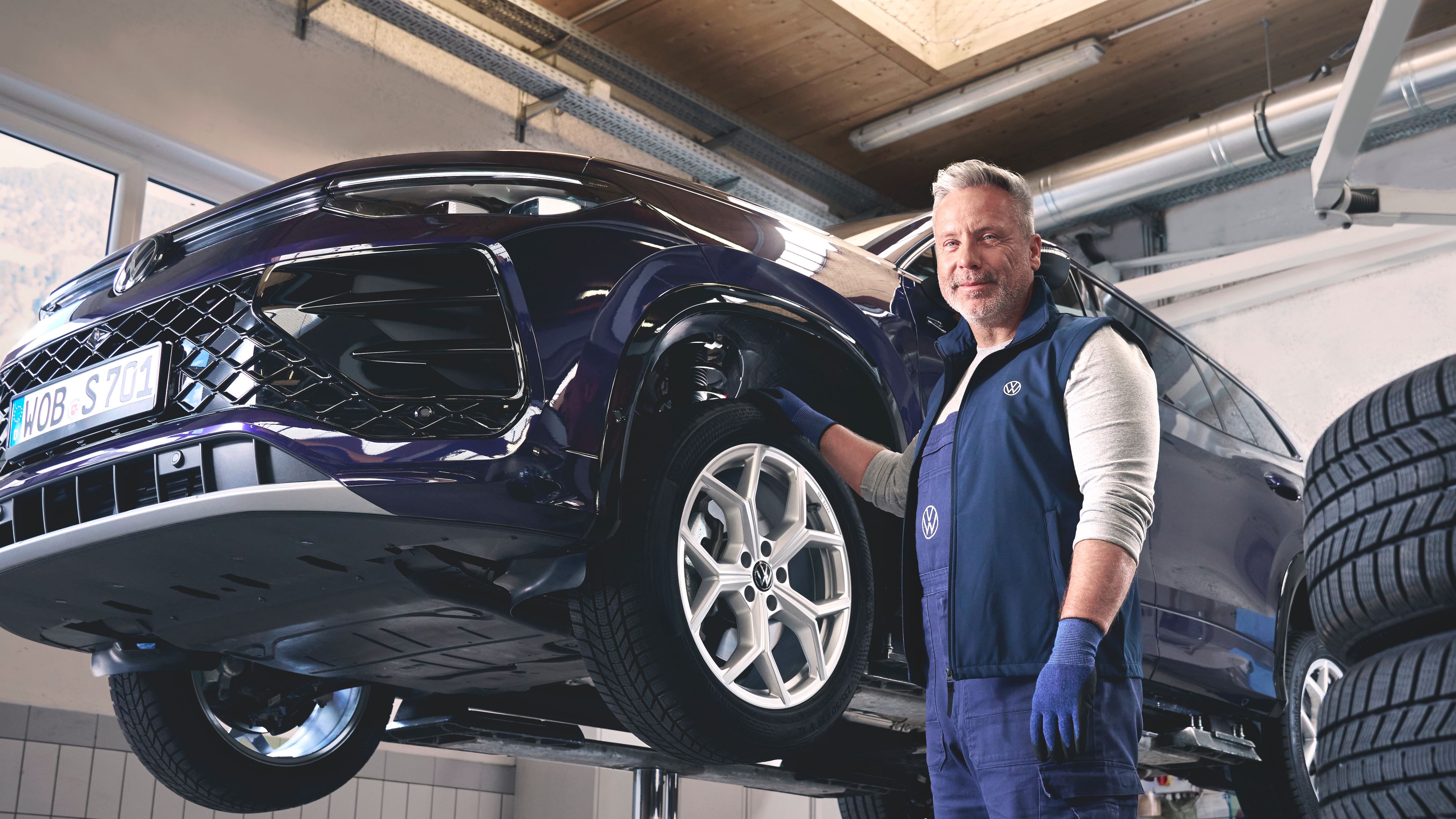Avtomehanik stoji ob dvignjenem avtomobilu v servisni delavnici Volkswagen. Okoli njega so kupi pnevmatik in različna orodja. Avtomobil je dvignjen za pregled ali popravilo.