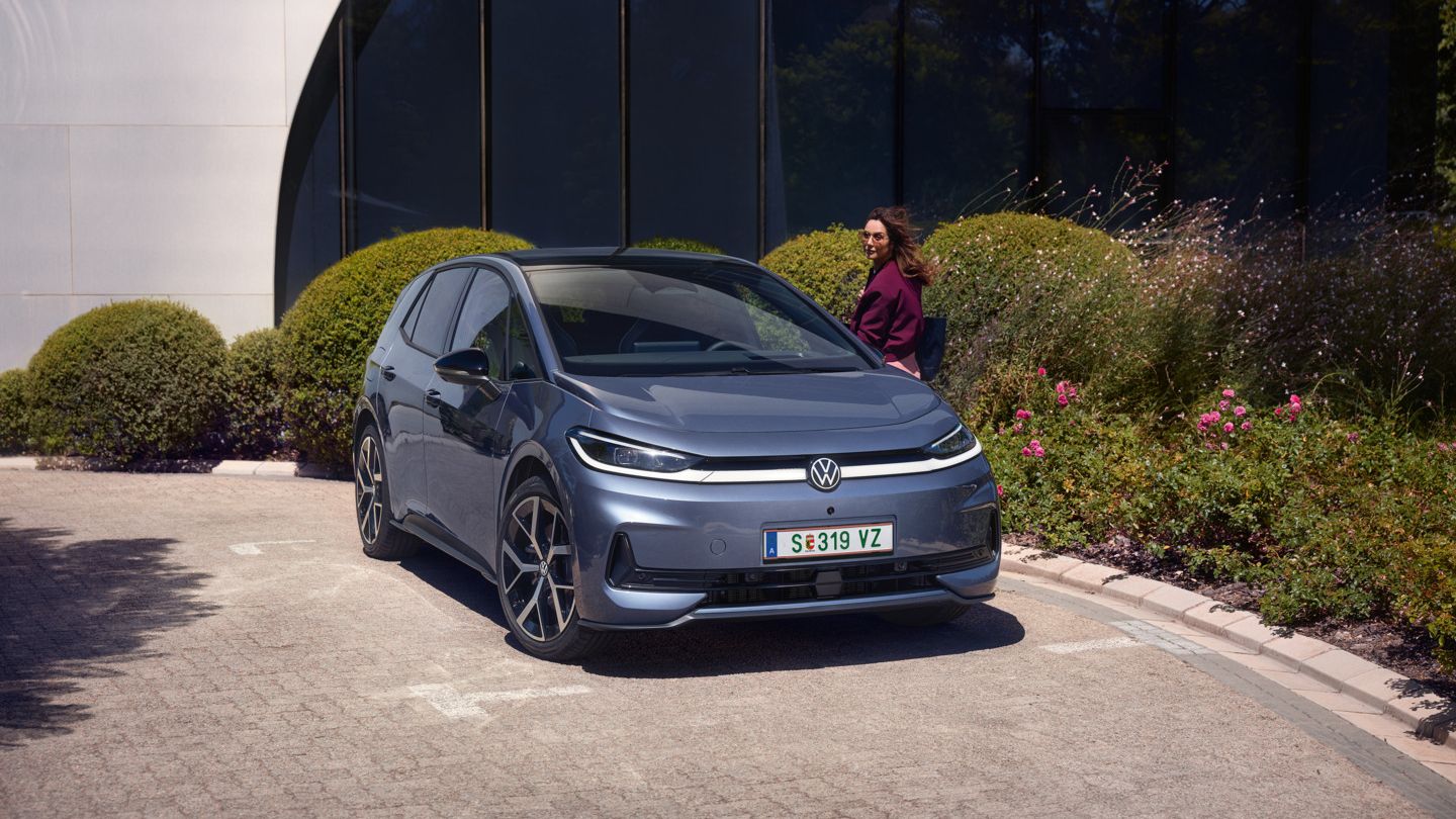 Ženska stoji pred voznikovimi vrati modrega VW ID.3 Neo, parkiranega pred sodobno pisarniško stavbo
