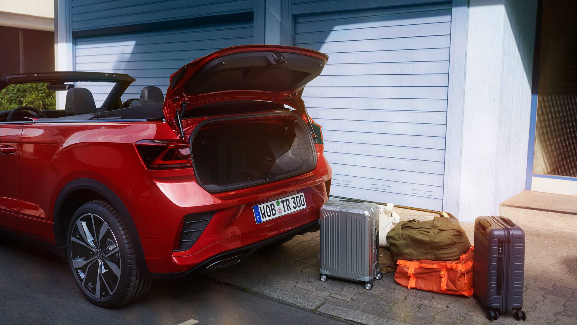 Rdeč VW T-Roc Cabriolet je parkiran ob robu ceste. Pogled s strani na odprt prtljažnik, poleg njega so dva kovčka in dve potovalki. 