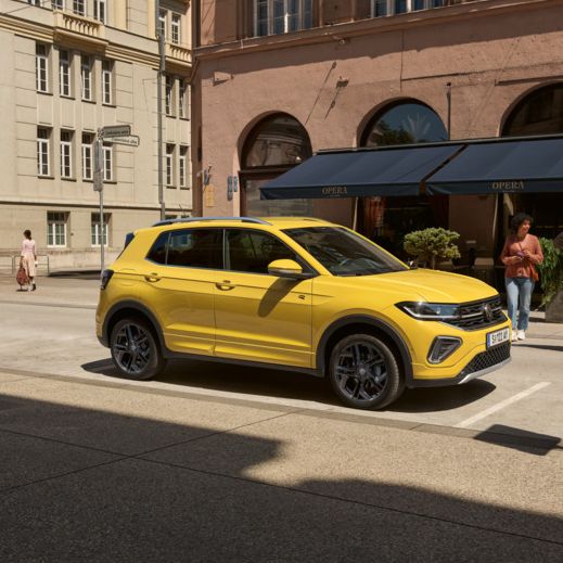 Ein gelber VW T-Cross ist in einer Stadt geparkt, hinter ihm sieht man Menschen