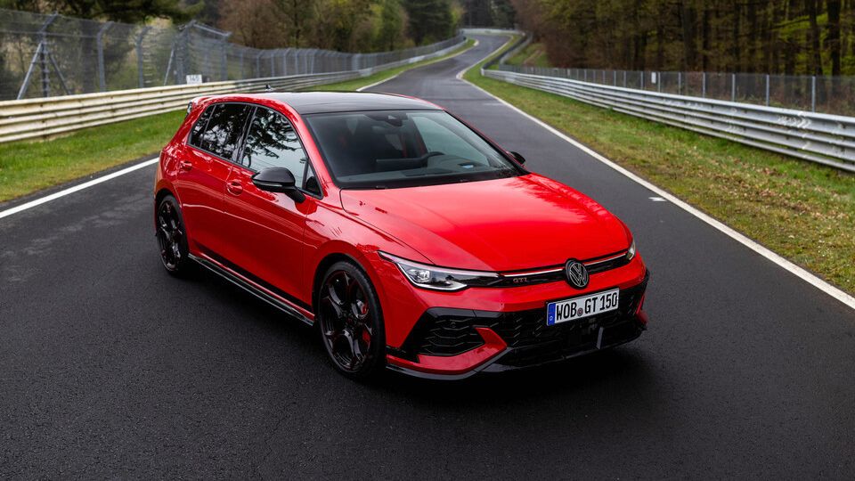 Rdeč športni avtomobil Volkswagen Golf GTI je parkiran na ovinkasti cesti med drevesi. Avto ima črna platišča in je označen z registrsko tablico. Okolica je gozdnata z zelenjem ob cesti.