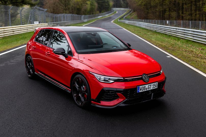 Rdeč športni avtomobil Volkswagen Golf GTI je parkiran na ovinkasti cesti med drevesi. Avto ima črna platišča in je označen z registrsko tablico. Okolica je gozdnata z zelenjem ob cesti.