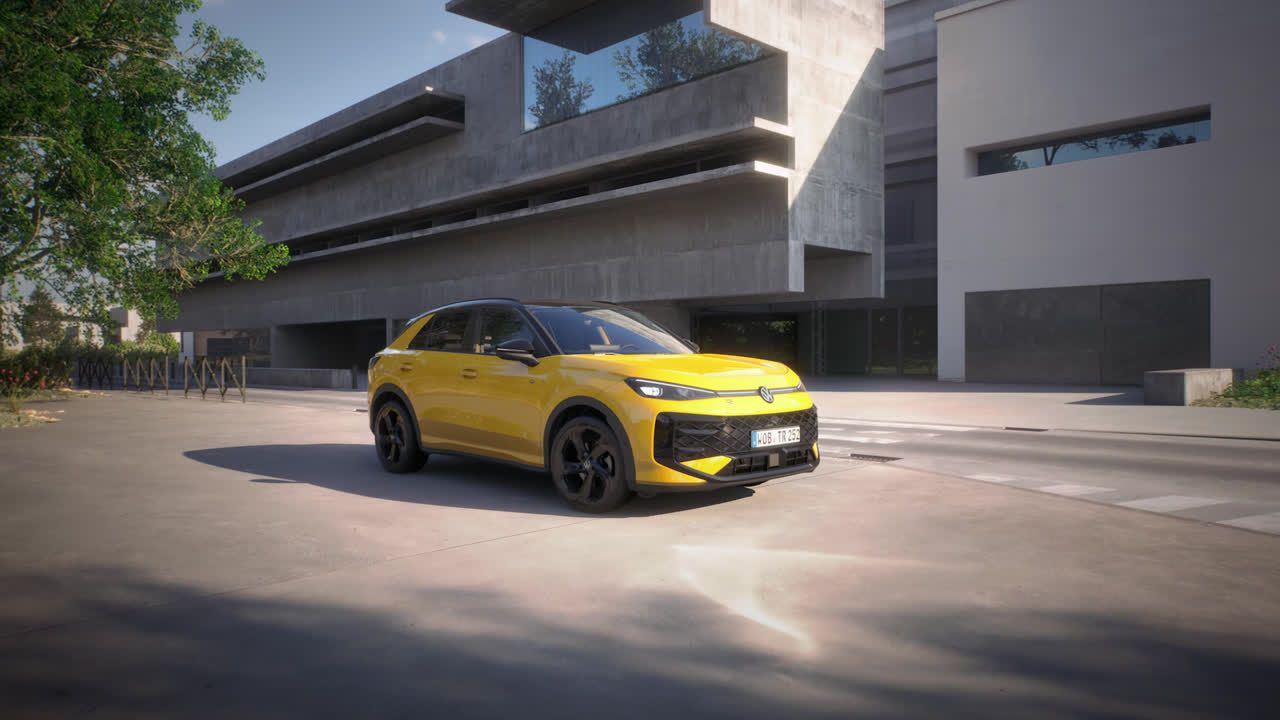 VW T-Roc rumene barve stoji parkiran na ulici pred moderno zgradbo iz betona in stekla. Vozilo ima črna platišča.